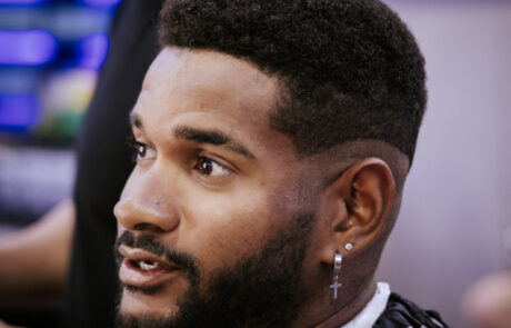 A person with short curly hair and a beard sits in a barber chair, wearing a striped cape. They have earrings and appear to be engaged in conversation.
