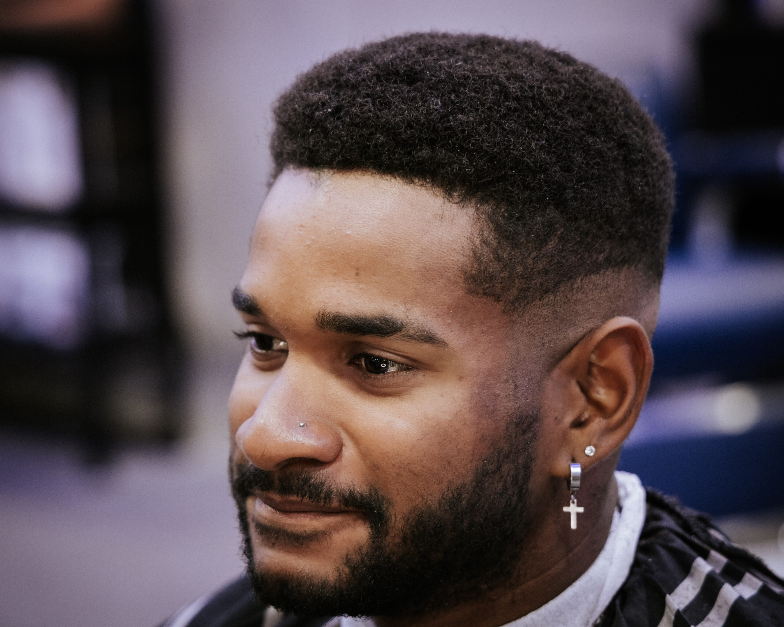 Young man with a neat beard and afro haircut, wearing a cross earring and nose stud, smiles softly. The background is blurred, conveying a calm atmosphere.