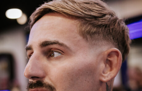 A man with a short haircut and a mustache looks to the side in a modern barbershop. The background features blurred lights and neon accents, creating a stylish, contemporary atmosphere.