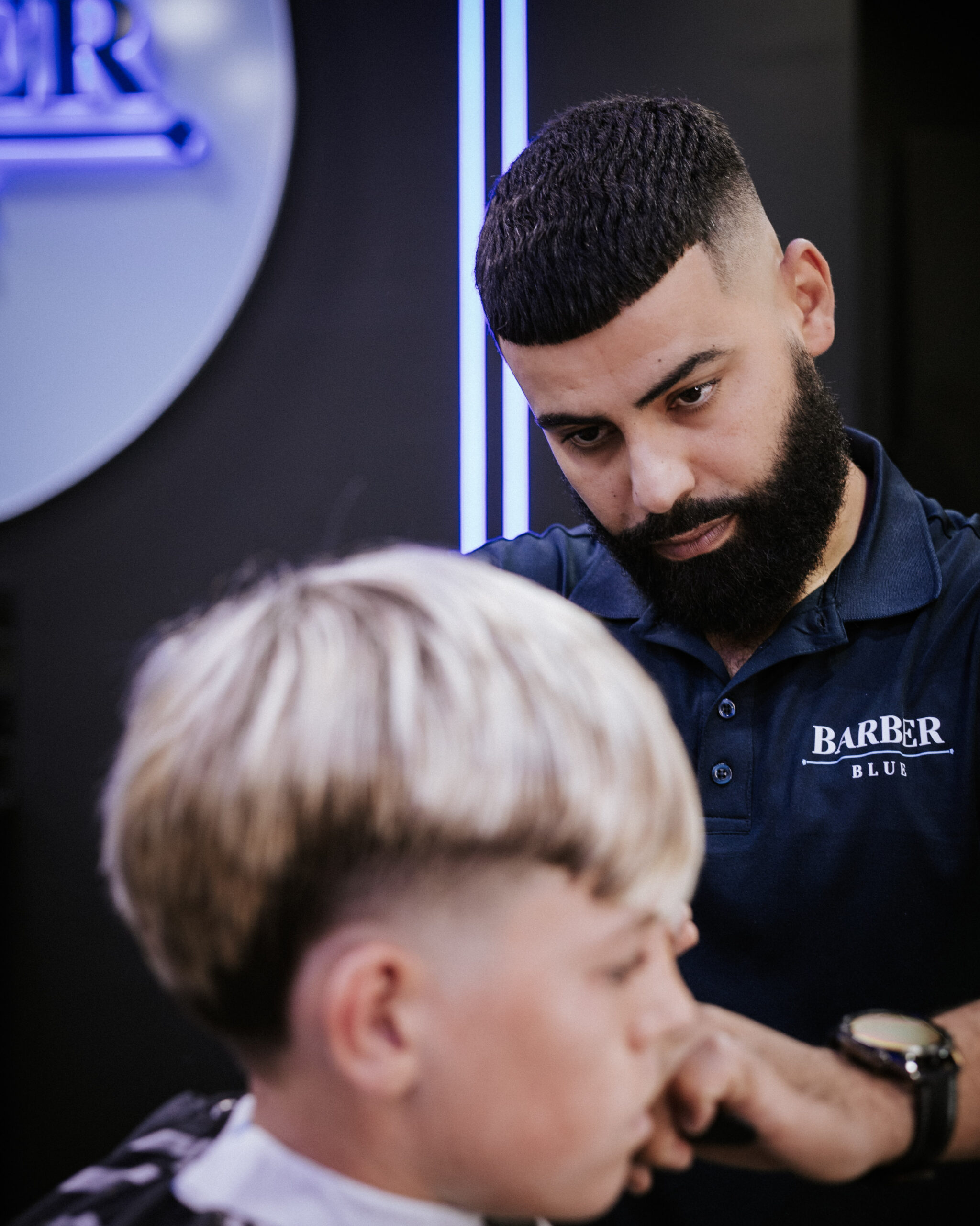 0T6A6701 (1) Barber attentively trims a young boy's blonde hair. The barber, focused and professional, wears a navy shirt with "Barber Blue" logo. Modern salon setting.