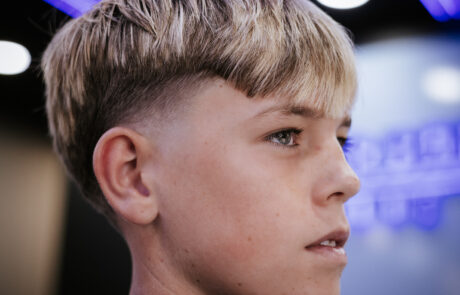 Profile of a boy with a modern short haircut, under cool blue lights. His expression is focused and thoughtful, with blurred background details.