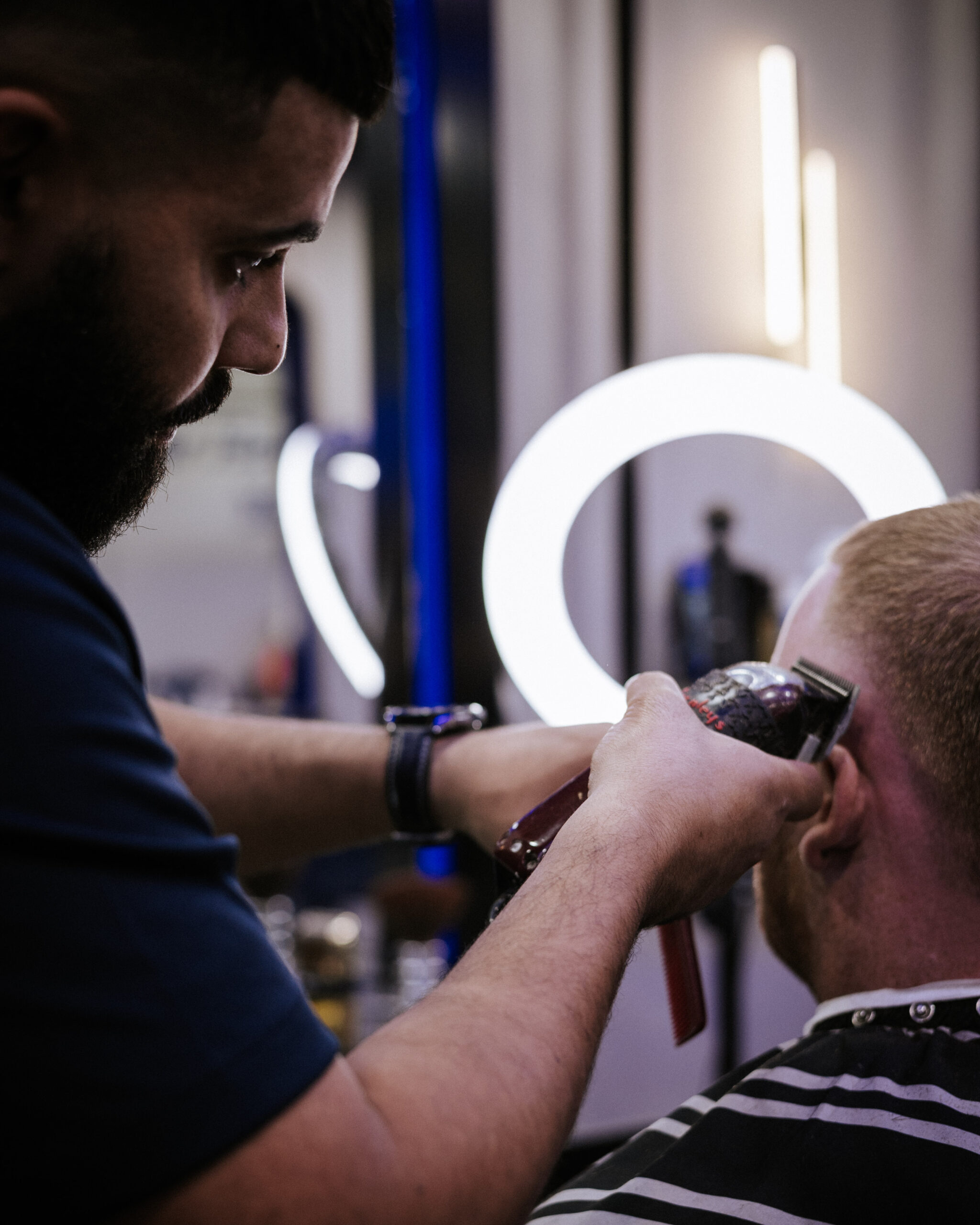 A barber uses clippers to trim the hair of a seated customer in a striped cape, with a circular light and mirror visible in the background.