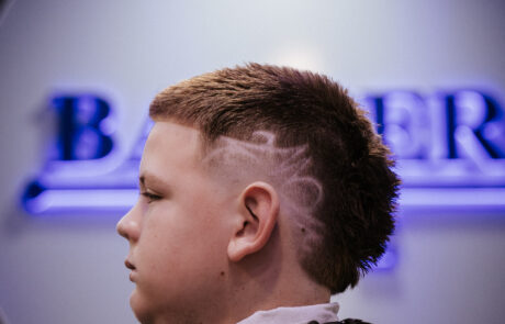 A young person with a short, styled haircut featuring geometric designs on the side sits in a barber chair. The background shows blurred barber signage.
