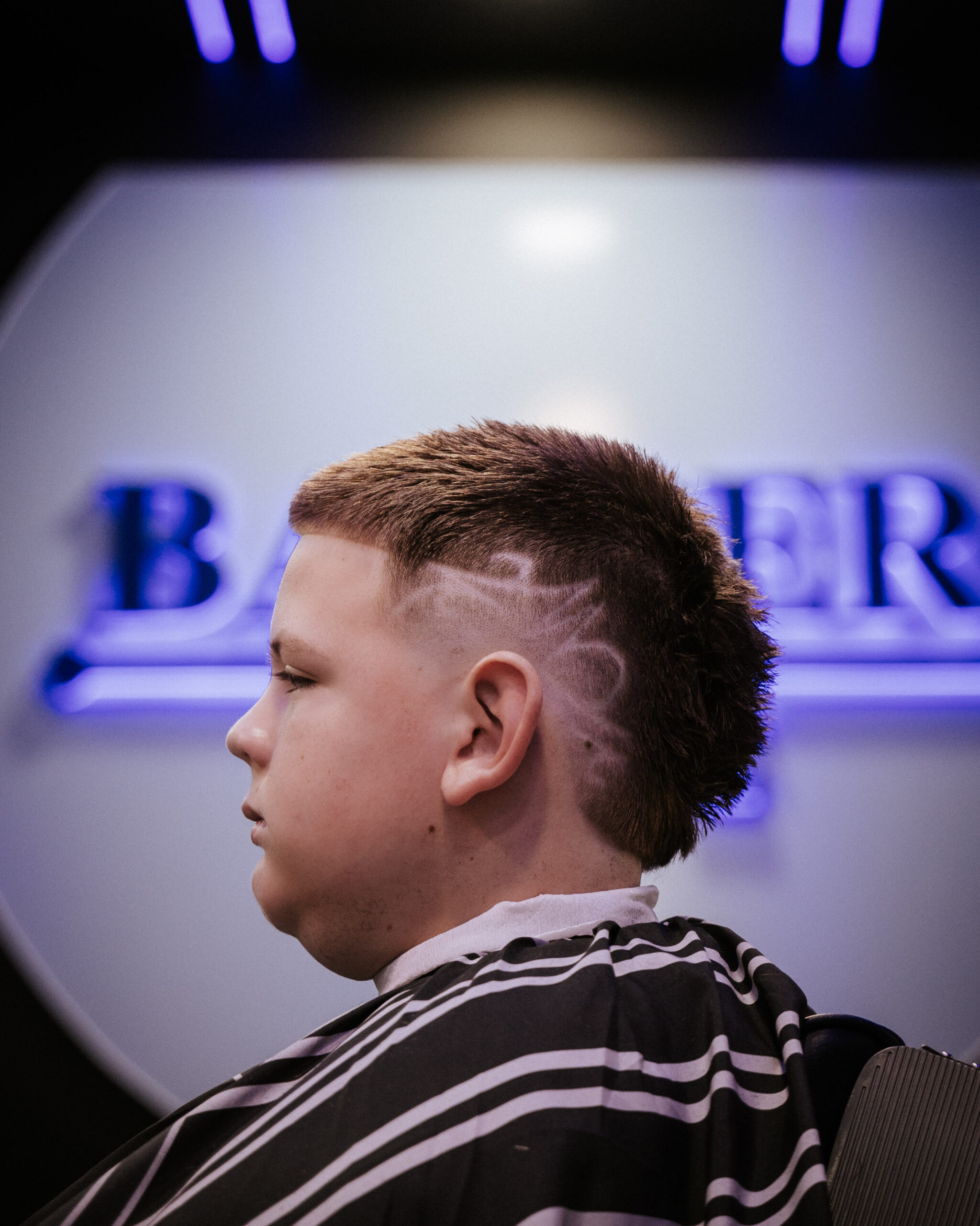 Profile of a young person with a stylish haircut featuring shaved designs on the side. They wear a barber cape. Background shows a blurred hair salon sign.