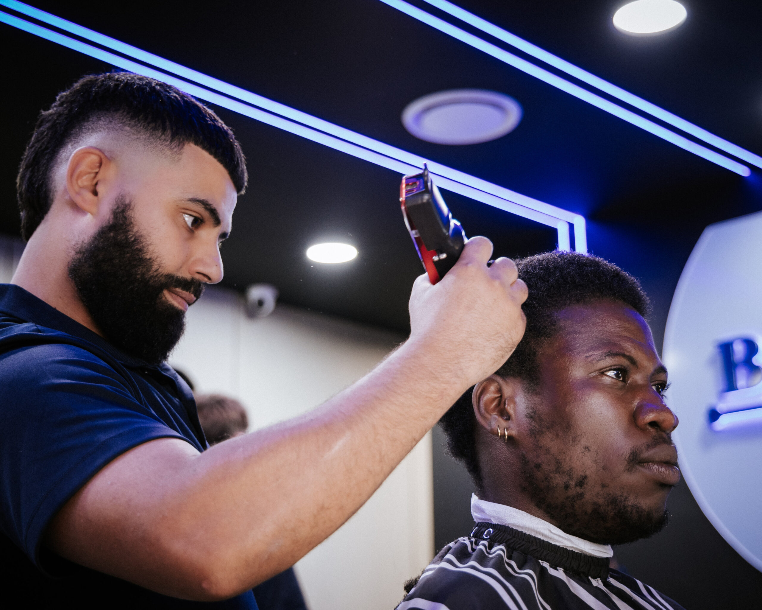 A barber styles hair with clippers in a modern salon featuring blue LED lighting and a chic atmosphere.
