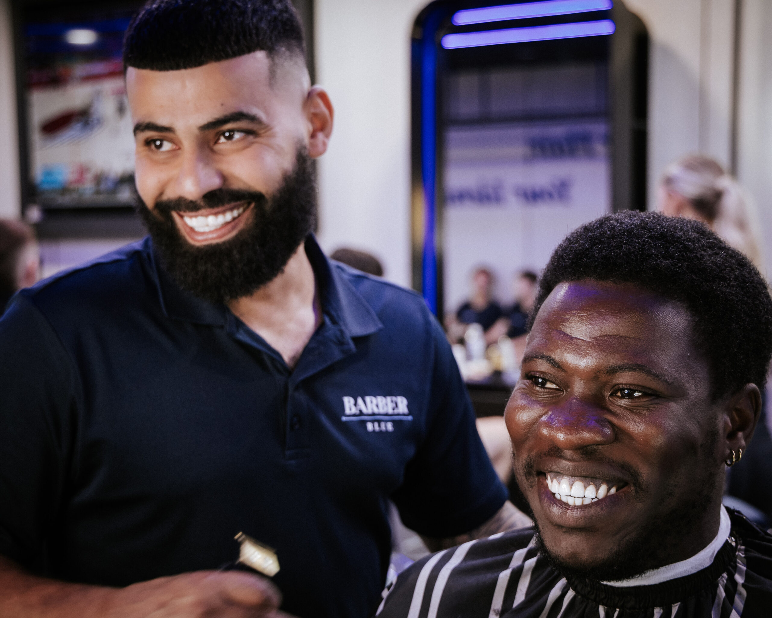 0T6A8172 Barber shop scene with a bearded barber in a blue shirt smiling at a customer with a striped cape. Both appear happy and relaxed.