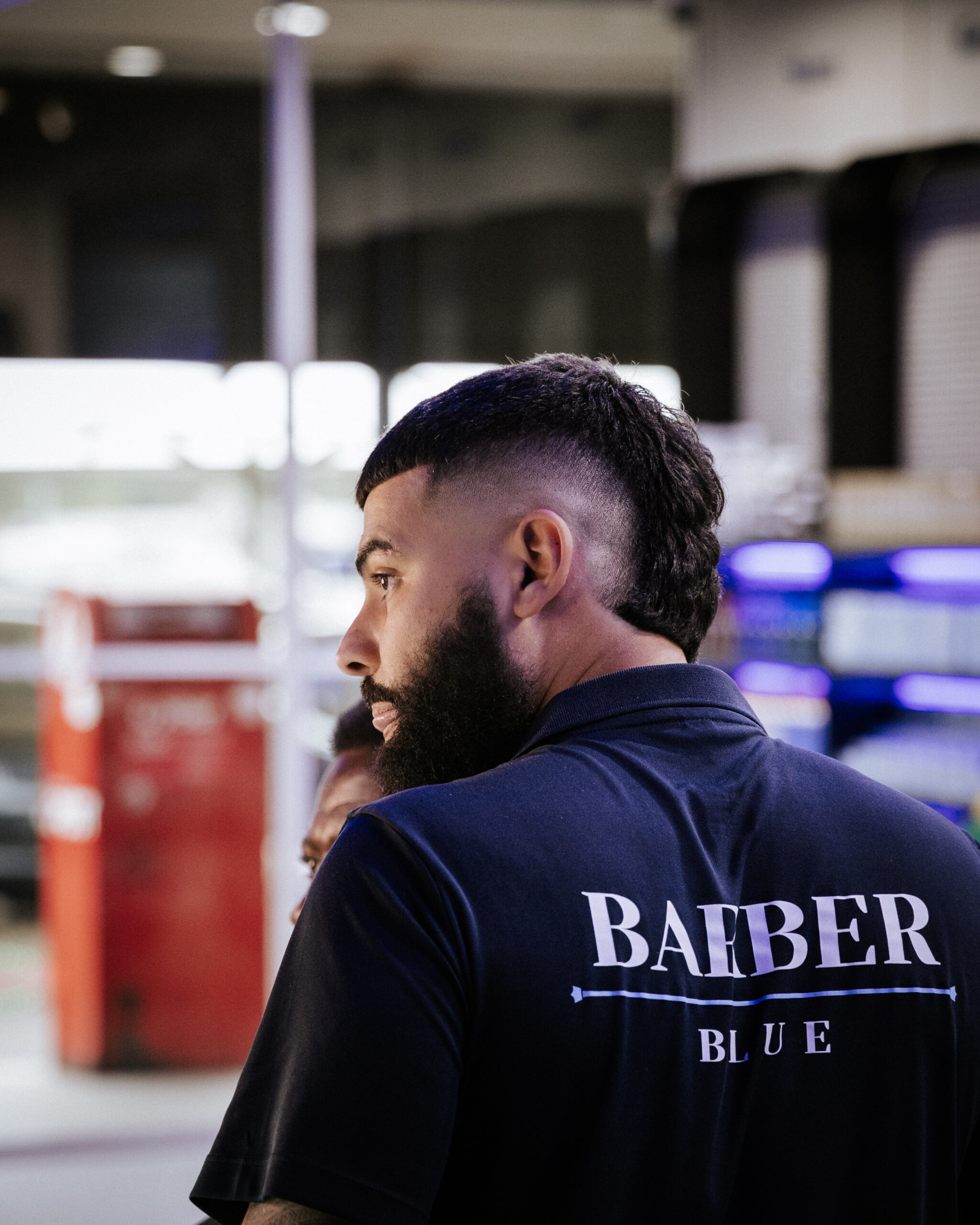 A man in a blue barber's shirt stands in a barber shop, showcasing a stylish haircut with a faded side view.
