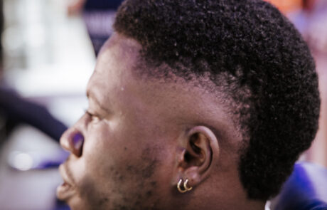 Profile of a person with short curly hair and fade haircut, wearing gold hoop earrings, looking focused. The background is blurred and colorful.