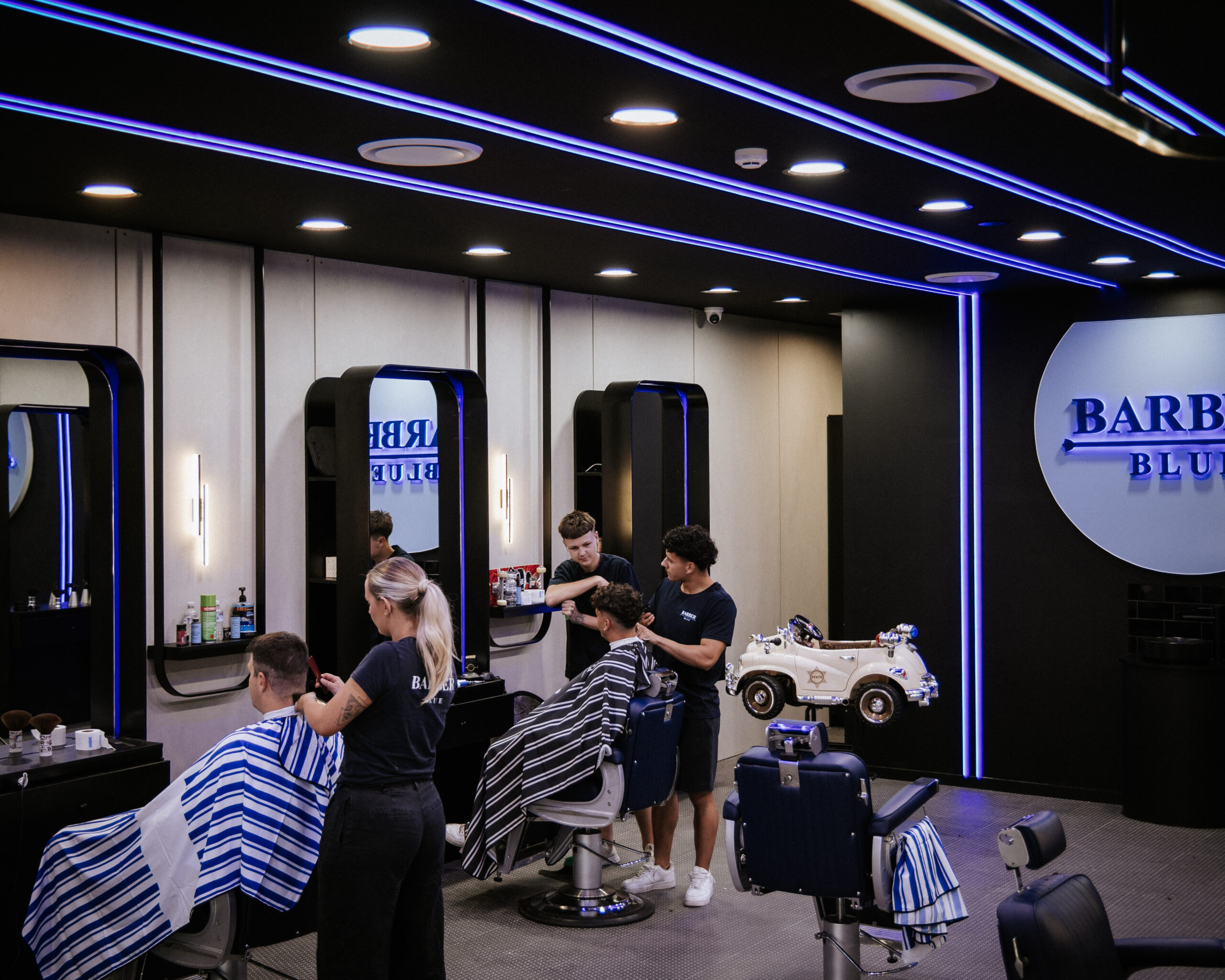 0T6A8657 Modern barbershop interior with dim lighting and blue neon accents. Two barbers give haircuts to clients covered in striped capes, creating a relaxed atmosphere.
