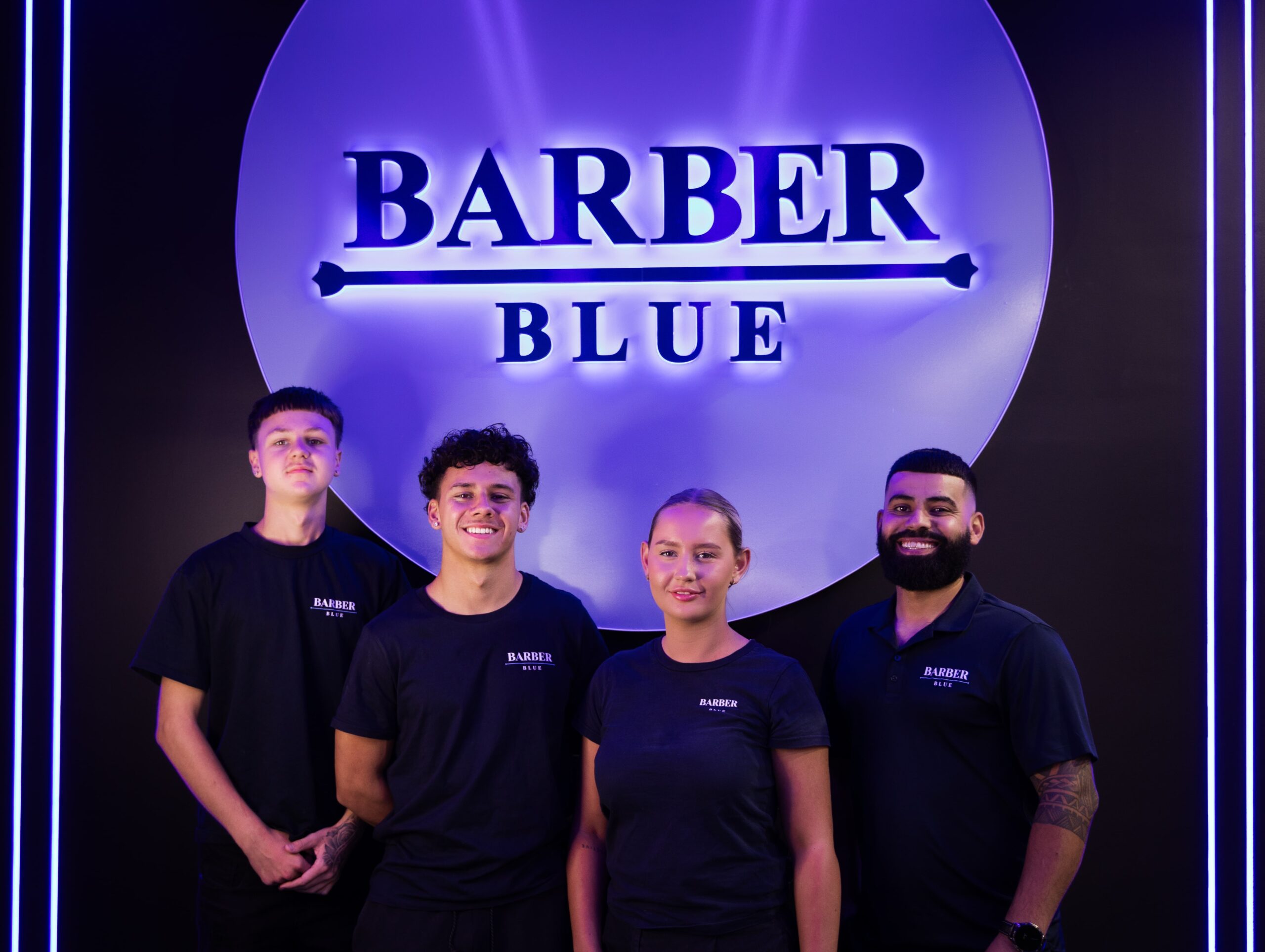 Four people in matching "Barber Blue" shirts stand smiling in front of a glowing blue and purple "Barber Blue" sign on a dark background.