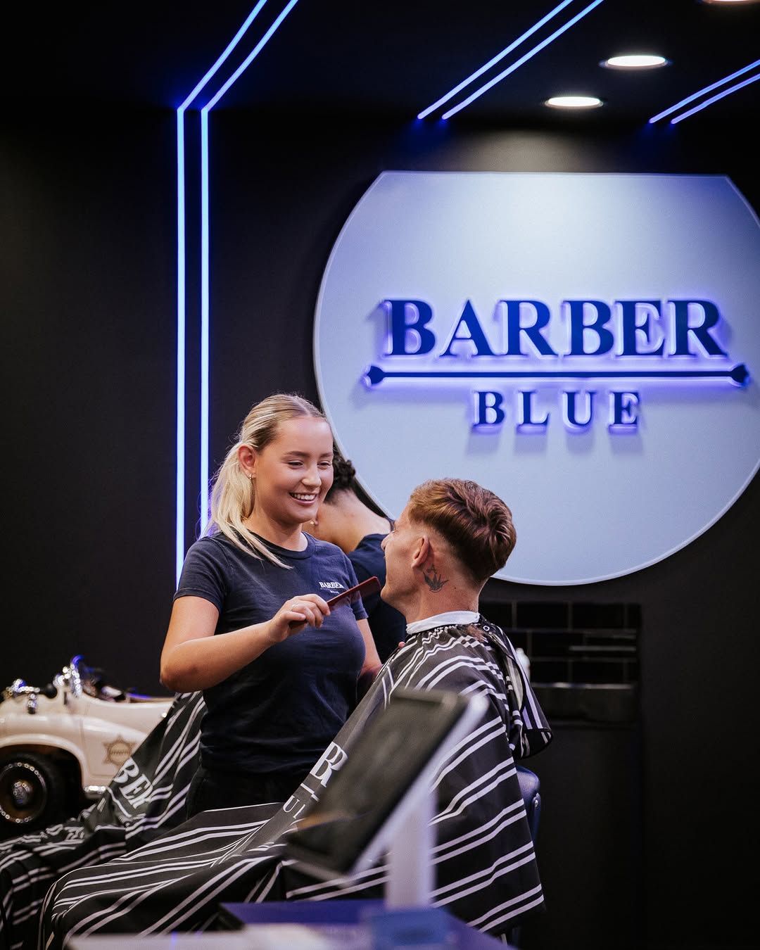 482335482_17867242371336843_2408436081204432383_n A smiling barber in a dark shirt combing a customer's hair at Barber Blue. The scene is lit with blue neon lights, creating a modern, friendly atmosphere.