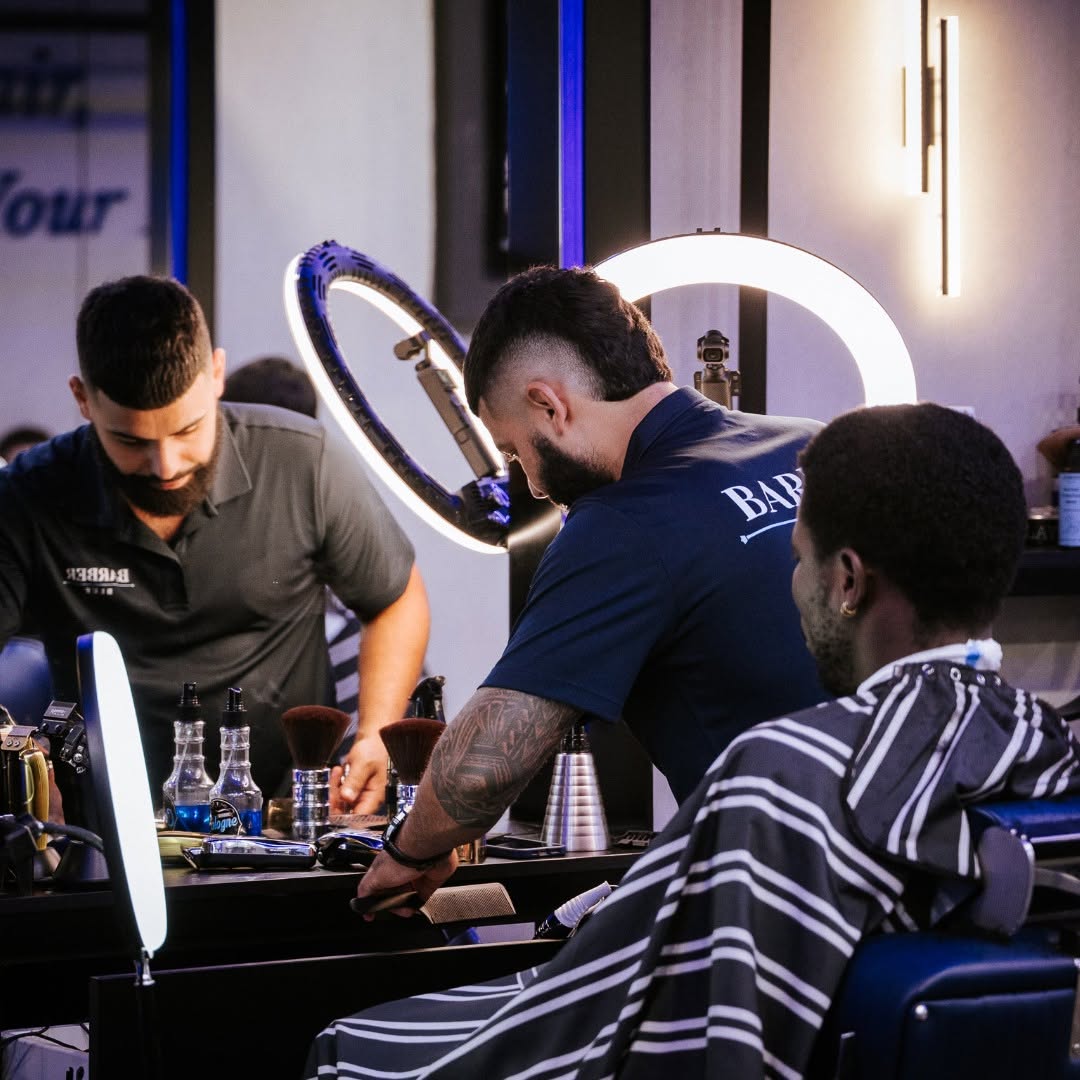485629799_17870252211336843_2359288209208660996_n Barber shop scene with two barbers, one styling a client's hair under bright ring lights. The client wears a striped cape, and tools are visible on the countertop.