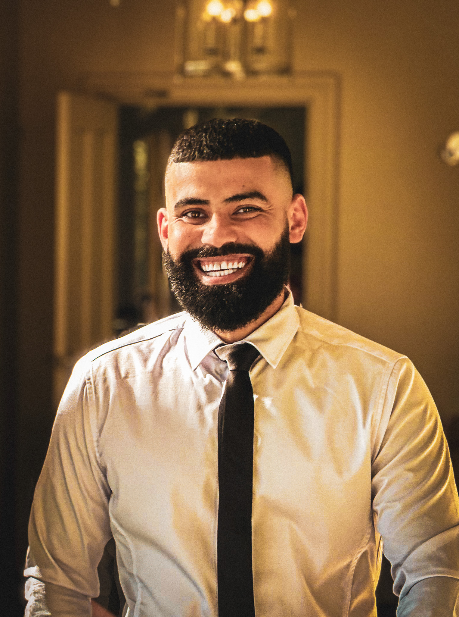 Smiling man with a beard stands in a warmly lit room, wearing a white shirt and black tie. The tone is joyful and inviting.